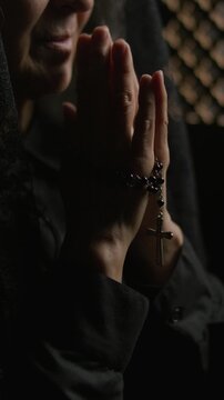 Close-up view of elderly woman holding rosary with cross in clasped hands and praying inside dim confessional booth. Vertical shot