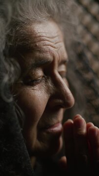 Devout senior woman praying deeply to God with eyes closed and hands pressed together inside confessional booth. Close-up view, vertical clip