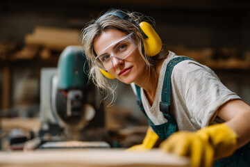 A female carpenter wearing protective goggles and ear muffs works attentively with wood in a workshop, demonstrating focus and skill in woodworking