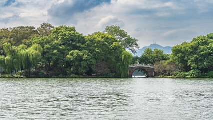 A picturesque urban lake. Ripples on the surface. Beautiful arched bridge over the water. Lush green vegetation on the shore. Blue sky, clouds. China. Hangzhou. West Lake Xi Hu 