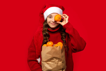 Playful girl in a Santa hat holds an orange in front of her eye