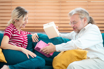 Smiling grandfather and granddaughter with gift boxes sitting on couch at home. Senior father with young woman indoors at home at Christmas, Holding present.