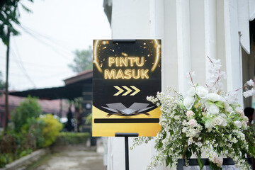 A black and gold directional sign in Indonesian reading "Pintu Masuk" (Entrance) at a formal event, flanked by elegant white floral decorations. Highlights event planning and arrival themes.