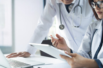 Two asian doctors discuss diagnosis, analyzing data on digital tablet in hospital office. Male and female medical professionals have a patient case conference, using tablet and laptop, close up
