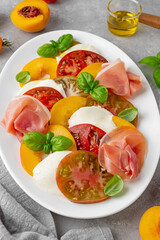 Salad of sliced multi-colored tomatoes, fresh peaches, mozzarella, prosciutto and basil on a gray concrete background. Delicious summer salad.