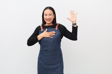 Asian adult woman wearing blue apron on white background swears with hand on chest and open palm raised upward, making oath of loyalty.