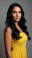 Naklejka premium Beautiful young hispanic woman with long wavy dark hair in yellow dress posing on grey background