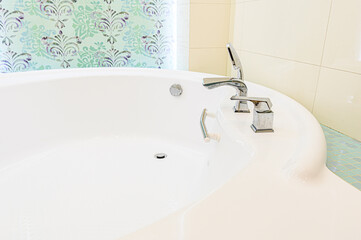 A close-up of a pristine white bathtub with chrome fixtures.  The background shows patterned wallpaper and neutral tiles