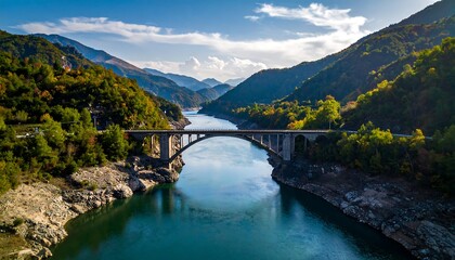 A concrete bridge spans a tranquil river, surrounded by lush, forested mountains under a bright blue sky, showcasing the beauty of the landscape