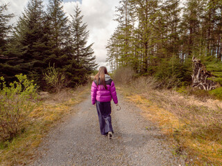 Obraz premium A woman in a purple jacket and dark pants walks on a dirt narrow road in a forest park. The road is surrounded by trees and the sky is cloudy. Day out in nature to get fresh air. Travel and tourism