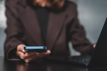 Businesswoman using smartphone and laptop for multitasking in workplace, technology and communication concept with shallow depth of field blur in background.