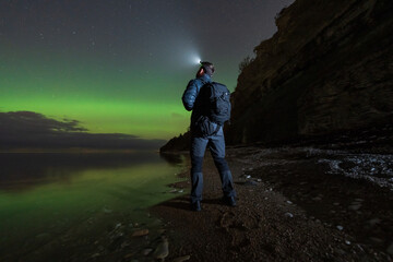 An adventurous man with a headlamp watches the magnificent green aurora borealis on the coast of...