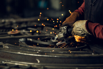 A worker wearing protective gloves uses an angle grinder on a metal workpiece, creating a dramatic spray of bright orange sparks in the dark workshop.