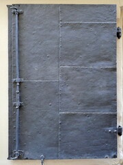 Sturdy historic metal door made of rectangular panels with rivets, dark surface and elaborate vertical locking mechanism and heavy hinges visible
