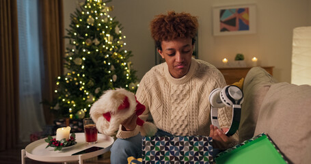 A smiling handsome man in a white sweater opens a gift box while sitting on a couch near a Christmas tree. He is curious, but his expression turns to disappointment and sadness because he doesn't like