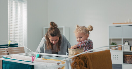 A mother hangs freshly washed clothes on the dryer in the bathroom while her daughter helps by...