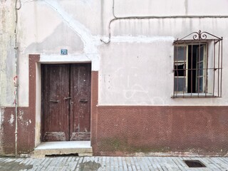 Old weathered wooden door and window with rusty bars on aged wall of abandoned house