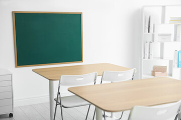 Blank green chalkboard, desks, chairs and shelving unit in classroom