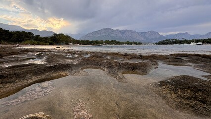 Tide pools on a rocky shoreline reflect the dramatic sunset sky over the Taurus Mountains in Kemer, Turkey. A moody and serene coastal landscape on the Mediterranean coast