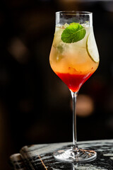 Layered cocktail in a wine glass with red and yellow gradient, ice cubes, lime slice, and mint garnish. Refreshing summer drink with elegant presentation on white background.