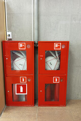 Two red fire cabinets with fire extinguishers and a fire hose against the wall. Ceramic tiles on the wall and floor.