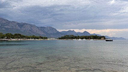 A moody, atmospheric view of Kemer's bay in Turkey, with yachts in the harbor and the massive Taurus Mountains under a dramatic, overcast sky. A beautiful coastal scene