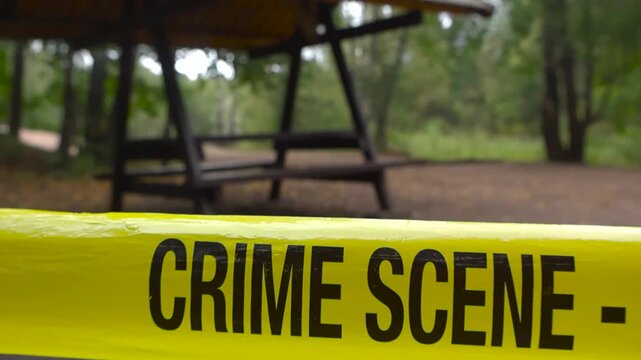 Yellow colored police crime scene tape in front of a grilling or a camping area at a cloudy day time forest during summer, with shallow depth of field and bokeh background.