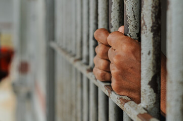 Obraz premium A person's hands tightly grasp the vertical, weathered metal bars of a rusty prison door or window, emphasizing feelings of confinement and despair in a stark, dimly lit setting.