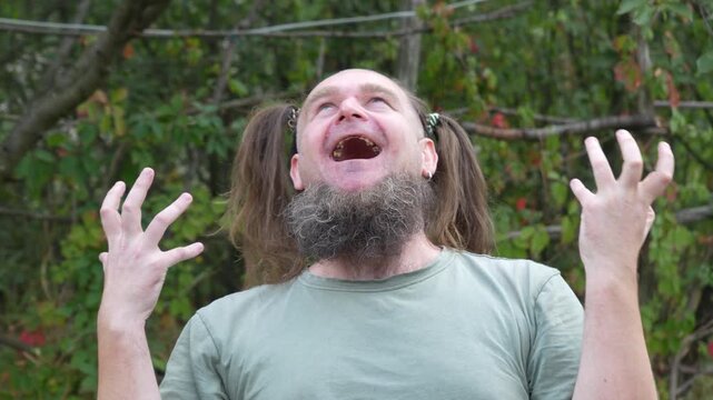 A bearded man with women's ponytails makes silly facial expressions and laughs in a humorous way during a staged outdoor shoot. Video captured in time-lapse at 50 fps.