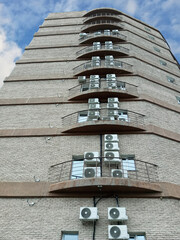 A brick high-rise building with windows and air conditioners. Against a cloudy sky, daytime. View from below.