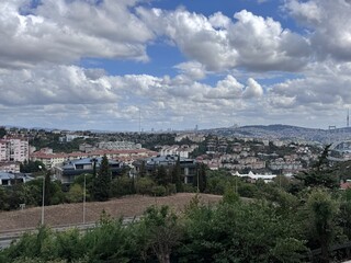 Obraz premium Panoramic cityscape of Istanbul with Bosphorus Bridge, residential buildings, green hills, and dramatic cloudy sky. Urban architecture and nature blend in a vibrant metropolitan landscape.