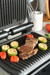 Open electric grill with beef steak, zucchini and tomatoes on table, closeup