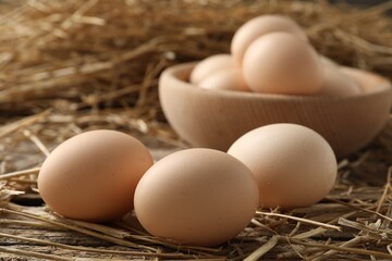 Raw chicken eggs in bowl and straw on wooden table, closeup