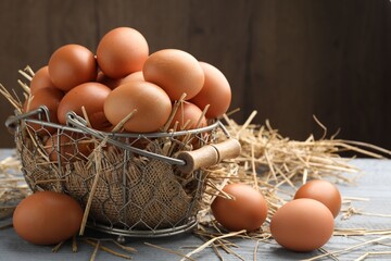 Raw chicken eggs, metal basket and straw on grey wooden table against brown background, closeup