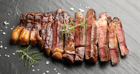 Pieces of delicious grilled beef steak with rosemary and salt on grey textured table, closeup