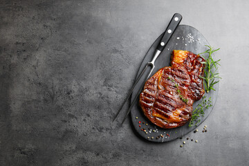 Delicious grilled beef steak with herbs, spices and carving fork on grey textured table, top view. Space for text