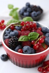 Different ripe berries and basil leaves in bowl on white table, closeup