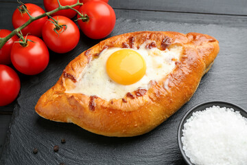 Tasty khachapuri with cheese, egg, tomatoes and spices on black wooden table, closeup