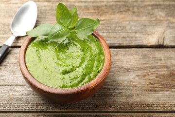 Tasty green pesto in bowl and basil leaves on wooden table, closeup. Space for text