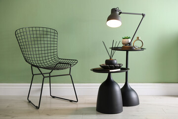 Stylish lamp, decor and reed diffuser on nesting tables near armchair against green wall indoors