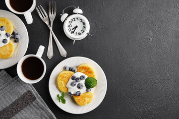 syrniki . Appetizing cheesecakes with sauce and frozen blueberries and cups of coffee on black table. Breakfast concept, top view