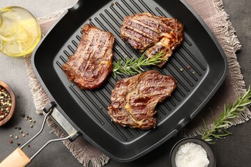 Pan with grilled beef steaks, rosemary, oil and spices on grey textured table, flat lay