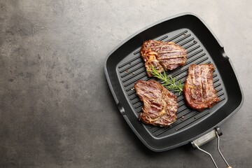 Pan with grilled beef steaks, rosemary and peppercorns on grey textured table, top view. Space for text