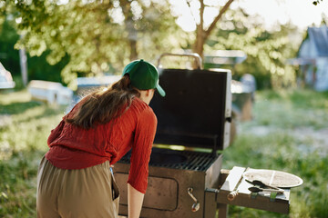 Young woman grilling outdoors, wearing a cap and casual clothing, surrounded by nature, evoking a...