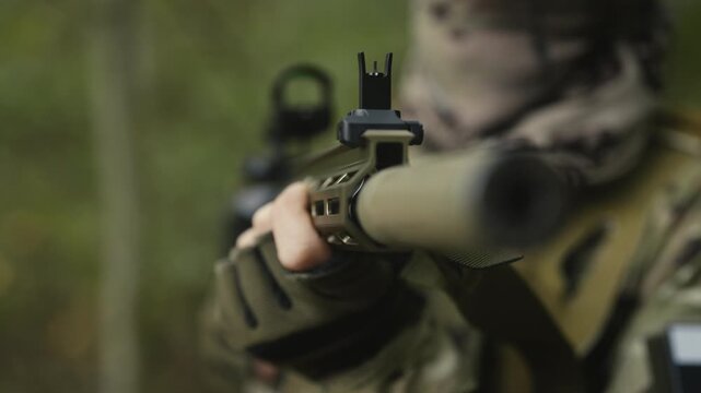 Camouflaged army soldier with AR15 carbine on a mission. Rifle with a collimator aiming at the camera. Selective focus