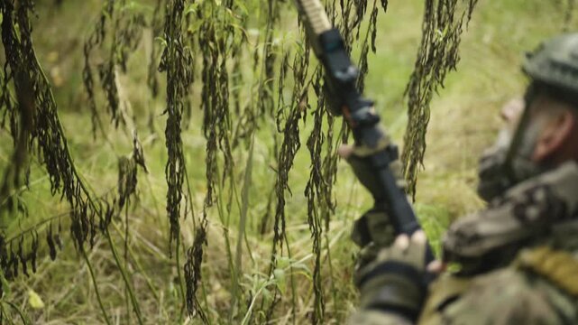 Camouflaged and masked army soldier with AR15 carbine with a collimator on a mission. Reloading the magazine hidden in a bush