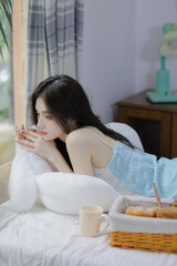 A young woman in a light blue lace dress relaxing by the window with a cup of tea.