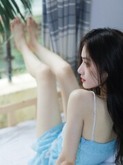 Woman in blue dress sitting on bed by the window with soft natural light.
