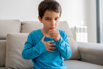 Child sitting on sofa at home and holding hand on chest. Male having asthma attack, difficulties with breathe, feeling severe pain or dyspnea