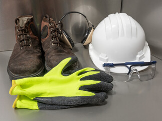 Personal protective workwear shoot on work location.The safety items are placed on metallic and includes a white hard hat, gloves, steel toe shoes, ear muff and goggles.A metallic in the background.